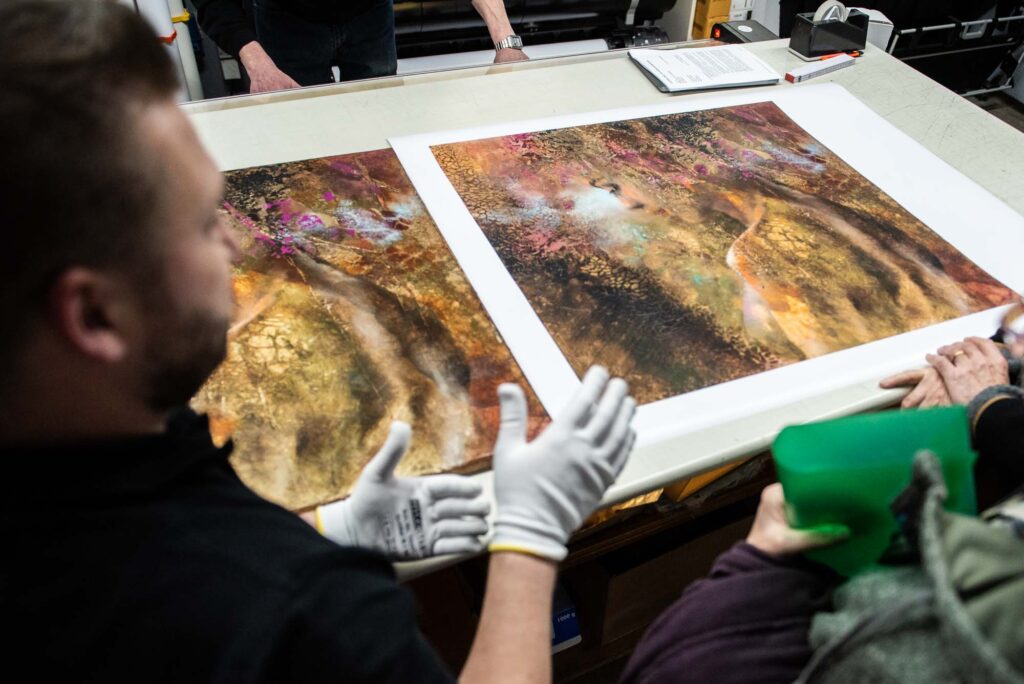 Ein Originalgemälde und ein gedrucktes Poster liegen zum Vergleich auf einem Tisch und werden von von Umstehenden betrachtet